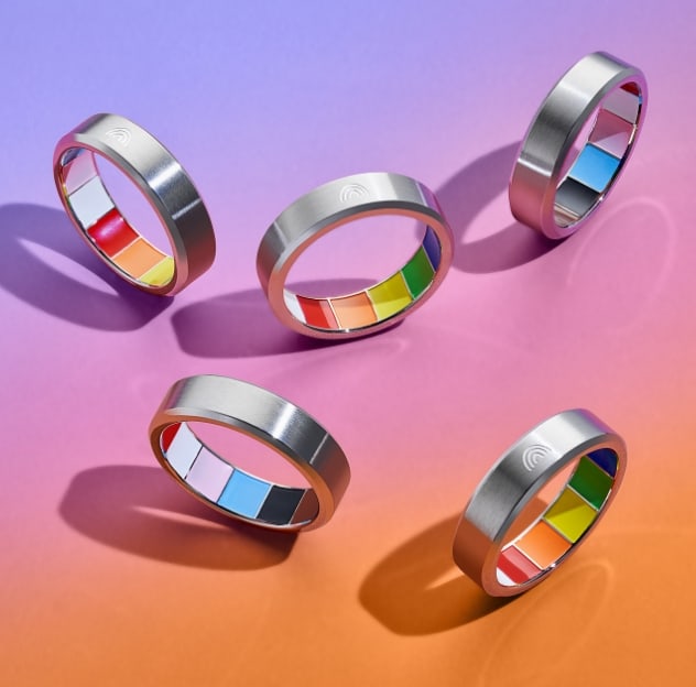 Five silver rings spin upright on their side, displaying a rainbow enamel interior that represents the colours of the Pride and Trans flags. The rings are set on a rainbow gradient background that transitions from light purple to pink to orange.