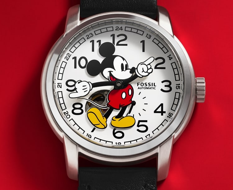 A classic watch with a white dial featuring Disney’s Mickey Mouse in his signature pose. He’s dressed in his iconic red shorts and yellow shoes. His gloved hands rotate to tell the time. The watch is set against a coordinating red background.