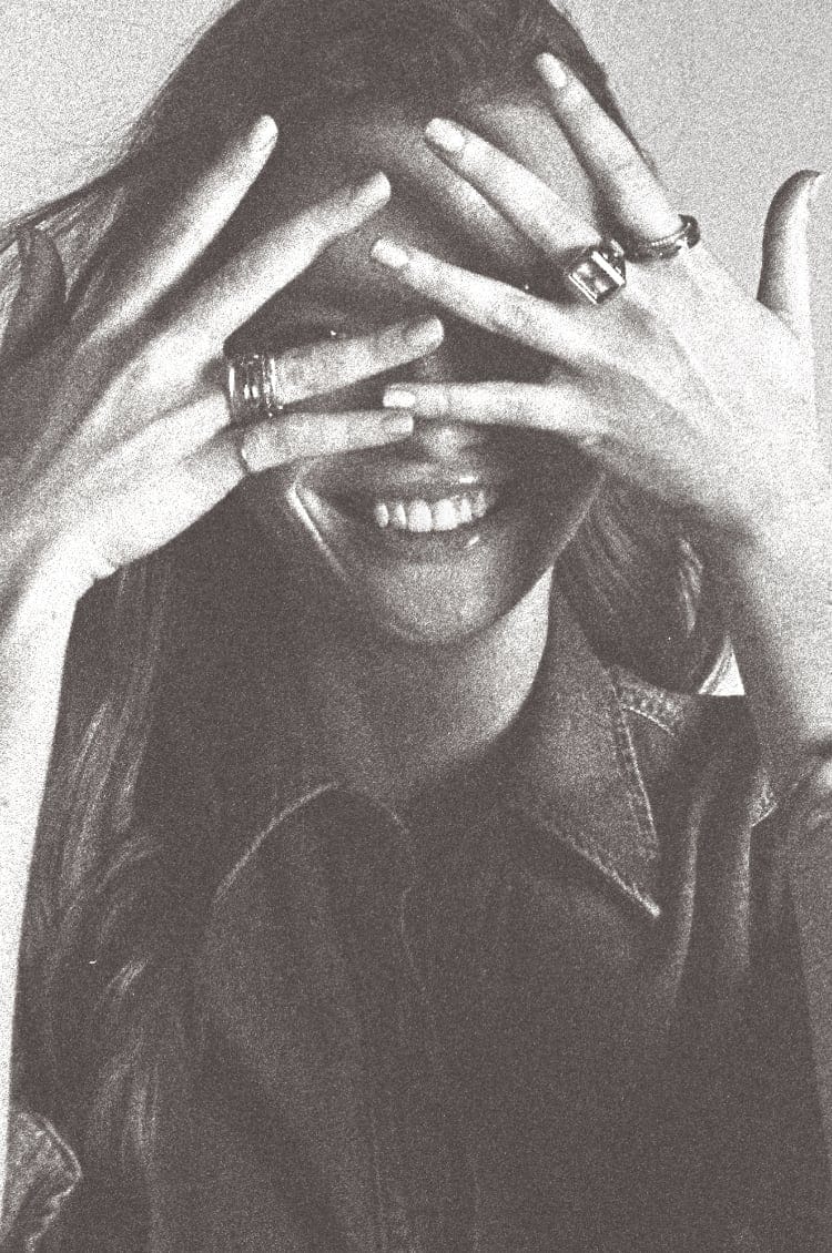 A black and white image of a woman covering her face with her hands while wearing a mix of Fossil rings.
