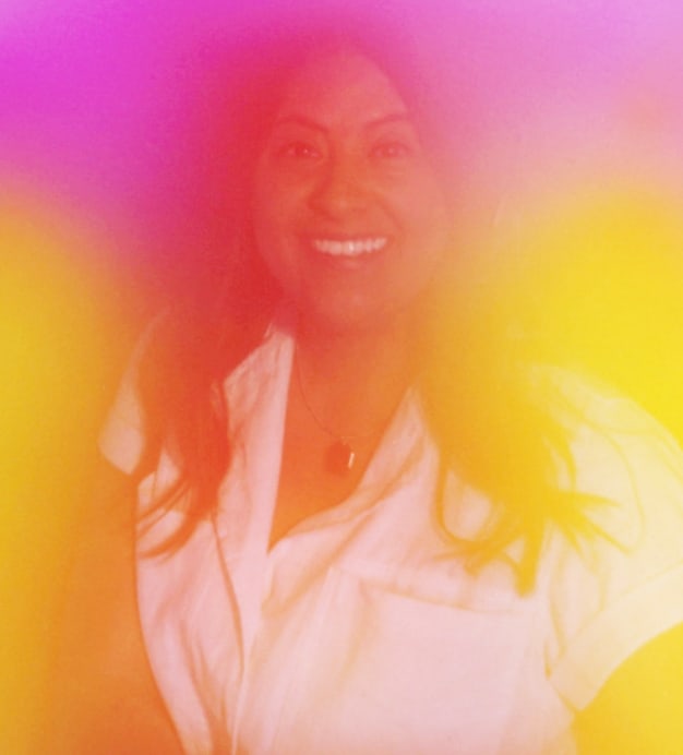 Die lächelnde Fossil Mitarbeitende Brenda, Product Manager für Herrenuhren. Über dem Foto liegt ein regenbogenfarbener Filter, der zeigt, dass alle mit Stolz ganz sie selbst sein können. Außerdem sieht man das Fossil PLUS Logo für LGBTQIA+ Mitarbeitende und Unterstützende mit zwei verschlungenen Regenbogen in den Farben der Pride- und Transgender-Flaggen.