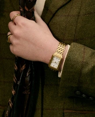 A woman's hand clutching a pebbled leather bag in ochre, wearing a Raquel watch with a gold-tone finish and black dial.