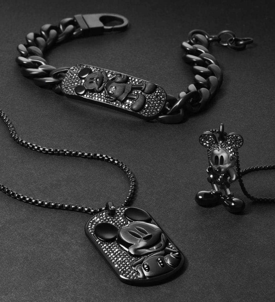 Tre gioielli di Topolino total black, ciascuno tempestato di cristalli di ematite. Un bracciale a catena con piastrina, una collana con piastrina e una collana con pendente a statuetta sono disposti in modo artistico su uno sfondo nero.