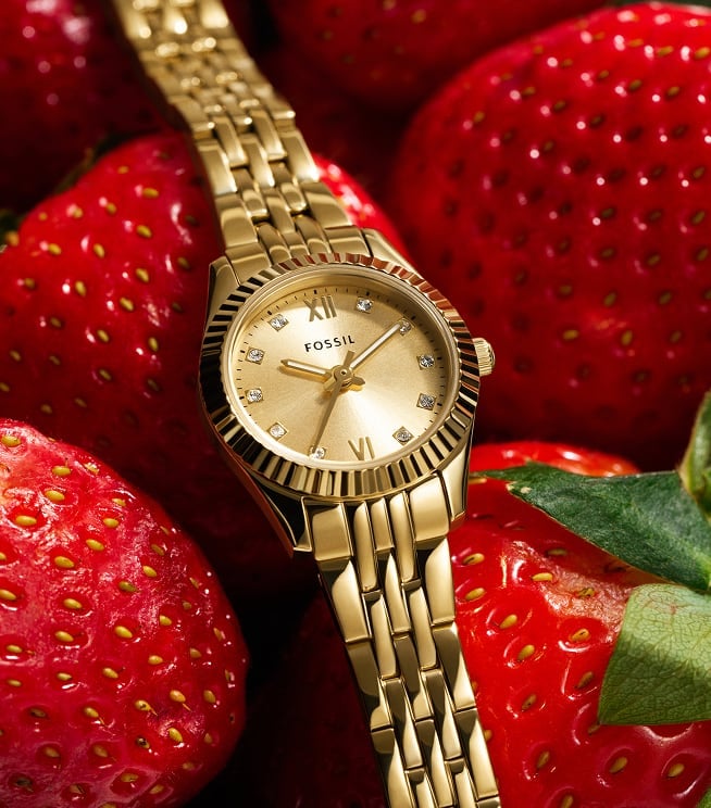 The Fossil Scarlette mini watch is placed atop fresh strawberries and worn by a woman picking up fruit at the market. Fossil fruit stickers add playful accents.