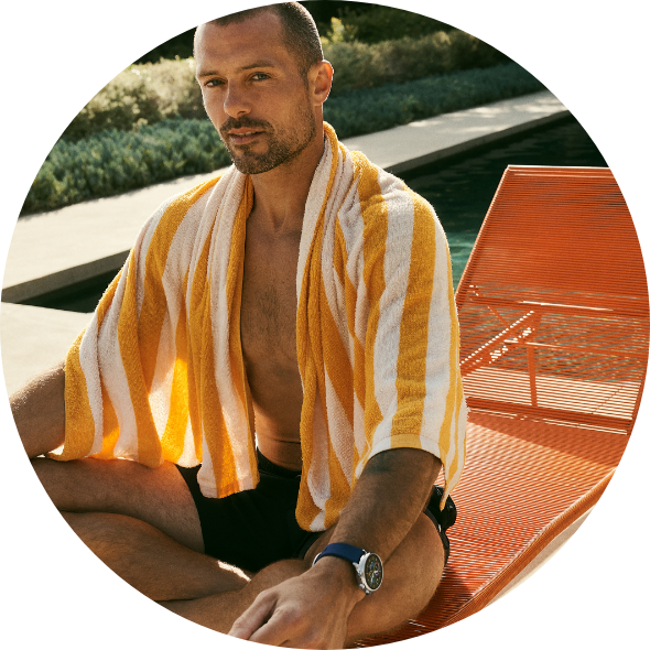 Gen 6 Wellness Editionスマートウォッチを身に着け、肩にストライプのタオルをかけ、プールチェアに座る男性。