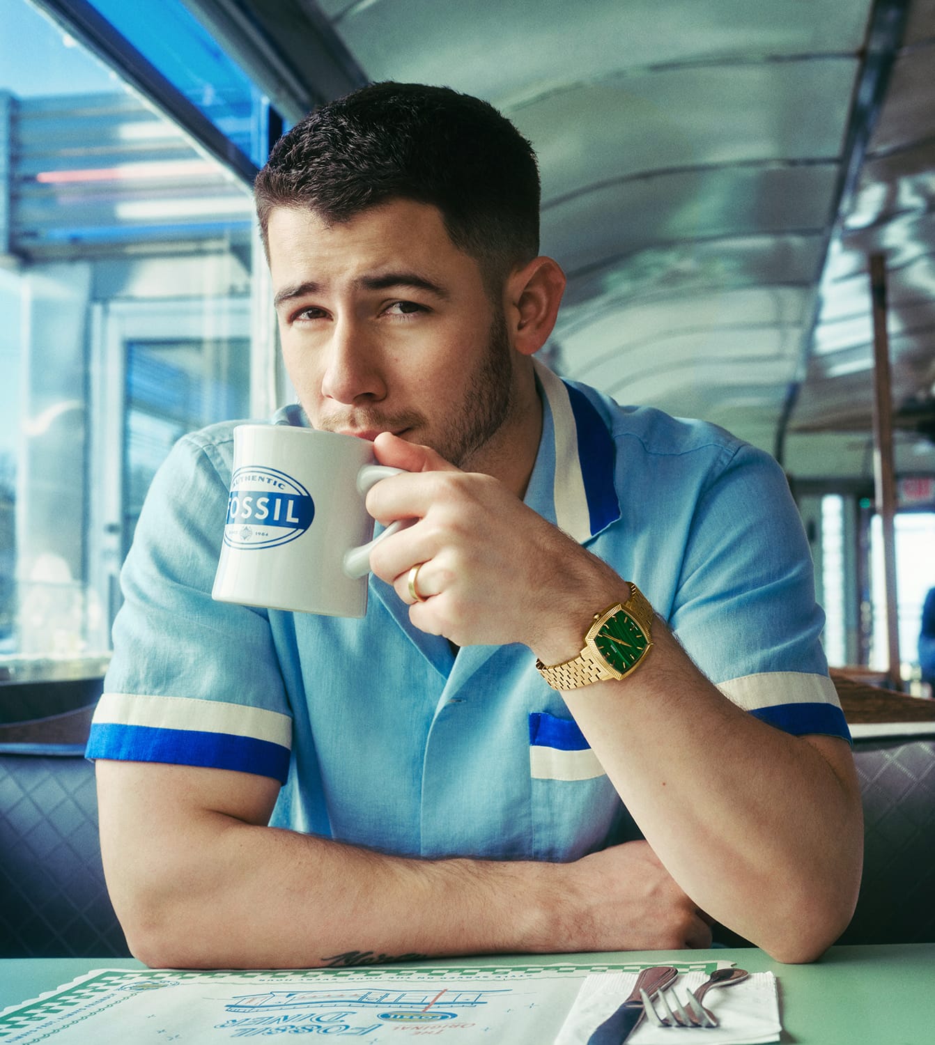Nick Jonas sitting in a diner, drinking from a Fossil coffee mug and wearing the gold-toned Machine Luxe watch with a green malachite dial.