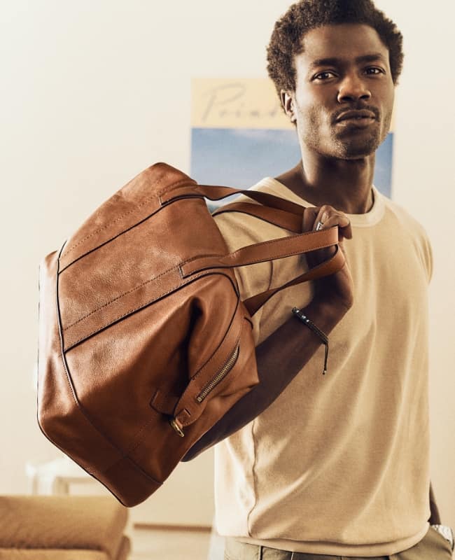 A man stands boldly with the Raeford duffle tossed over his shoulder. It has soft brown leather, a lightweight, convertible silhouette and a removable and adjustable crossbody or shoulder strap.
