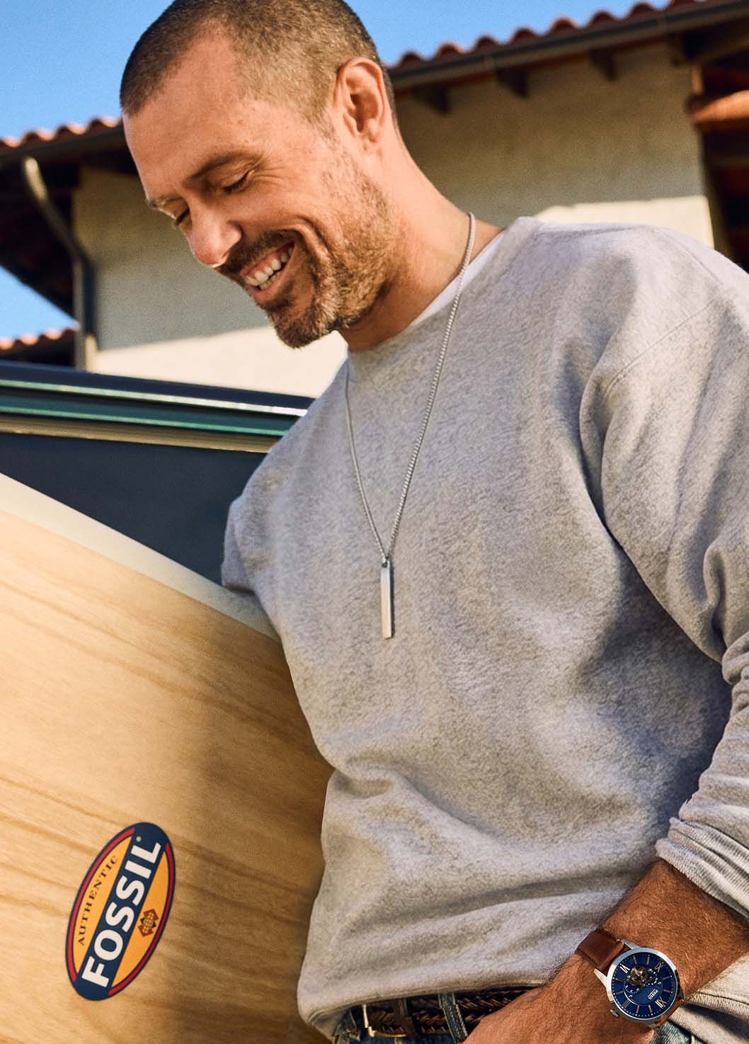 The stainless steel Fossil Ellis ID Necklace, shown in a lifestyle image of a man carrying a surfboard.