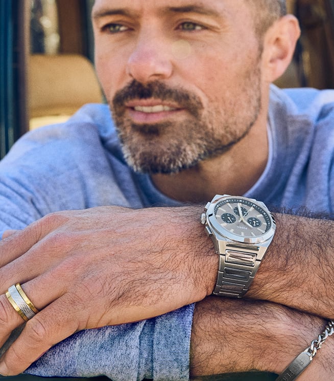 A man is looking out with his arms folded on the open car window. He's wearing a silver fossil ring, a silver Everett watch, and a silver ID bracelet.