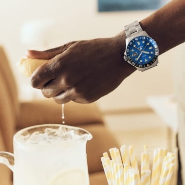 La mano de un hombre que exprime un limón en una jarra de limonada, y que lleva un GMT Fossil Blue de acero inoxidable con esfera azul. 
