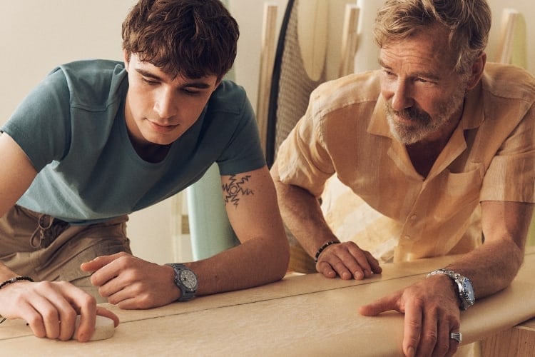 A father with his son sanding a surfboard while wearing Fossil products.