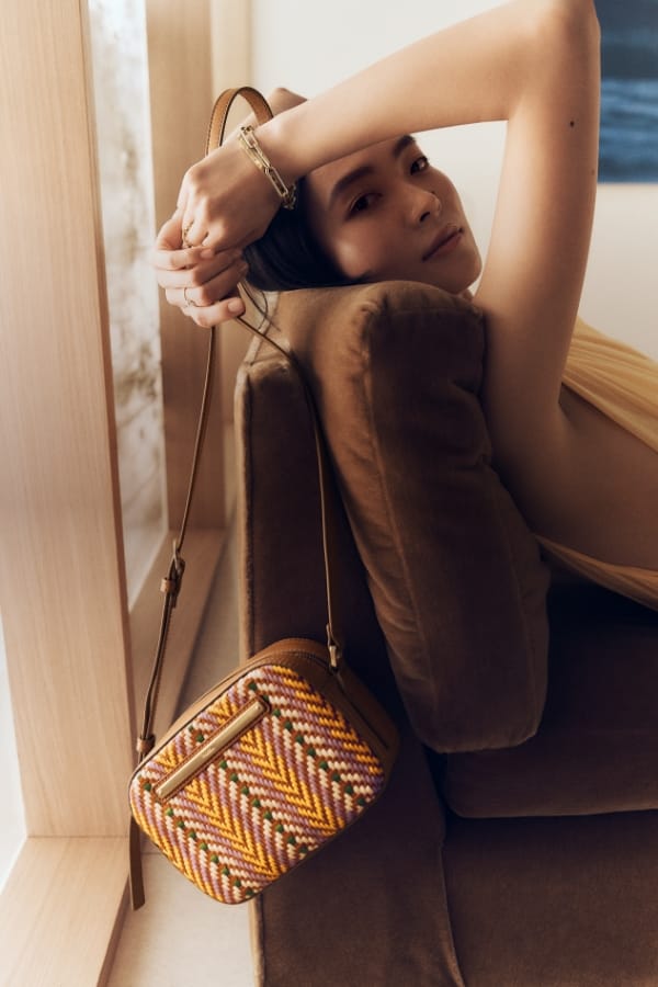 Eine auf einem braunen Sofa sitzende Frau, die den Riemen der bunten Kameratasche Liza hinter ihrem Kopf hält, sodass die Tasche hinter ihr drapiert ist. 
