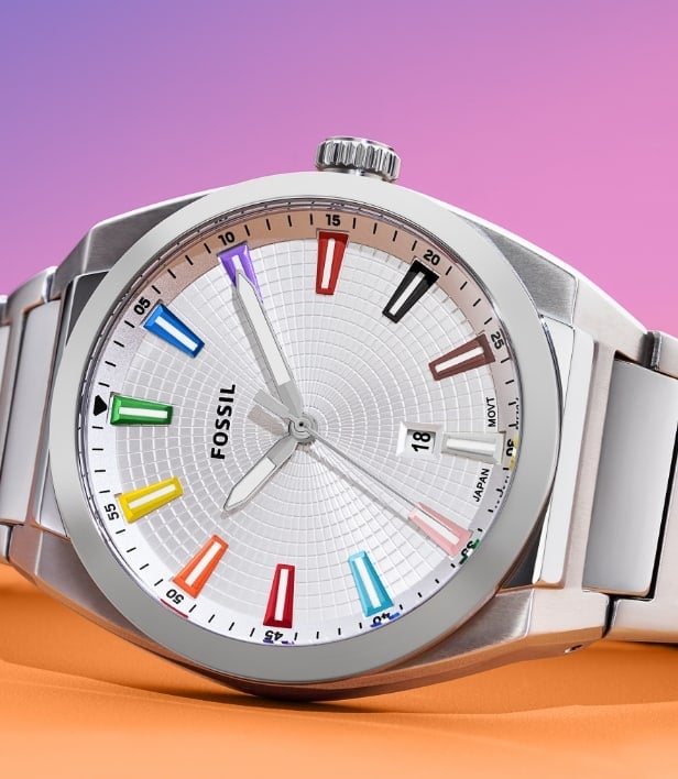 Un reloj plateado de género neutro con una esfera con textura e índices de arcoíris que representan los colores de las banderas del Orgullo y Trans. El reloj está colocado sobre un fondo degradado en los colores del arcoíris que pasa del morado claro al rosa y al naranja.