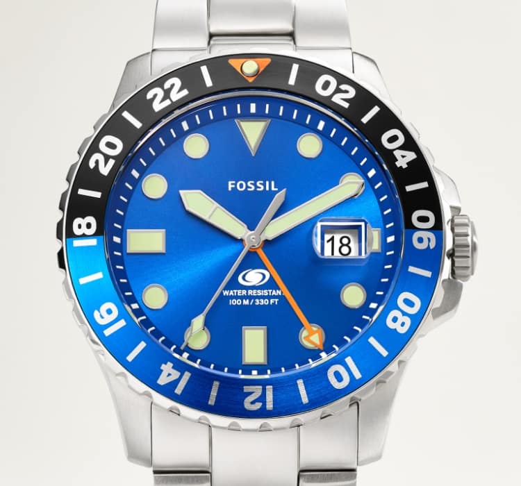 A man sits in a chair. He's wearing a Fossil Blue GMT watch: a stainless steel bracelet watch with a blue dial featuring geometric indices.