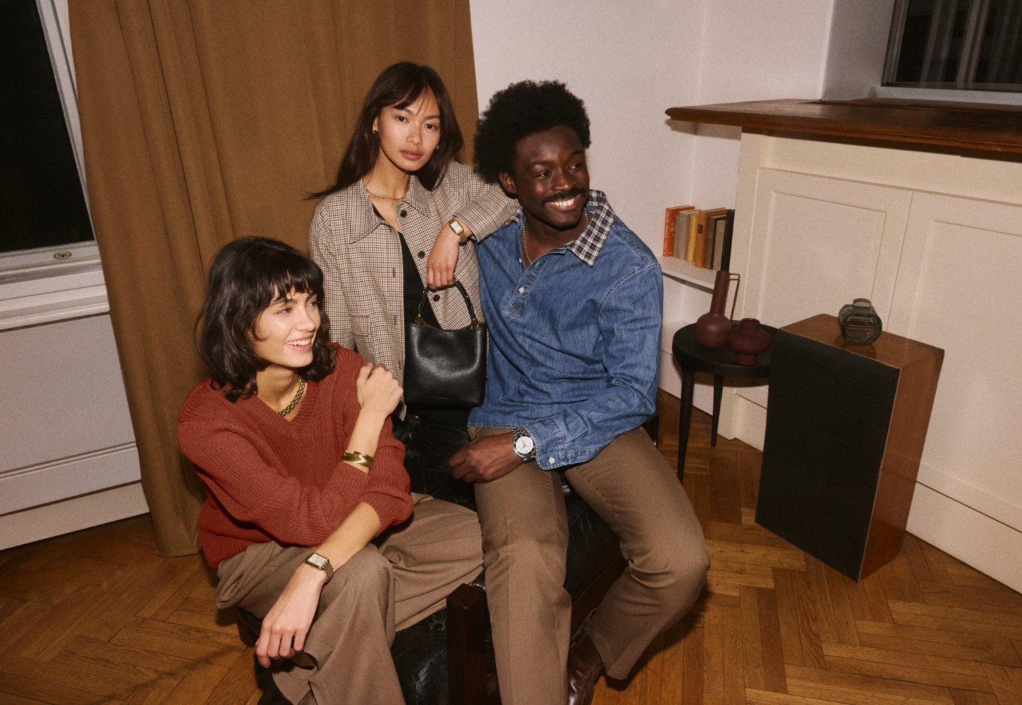 Two women sitting with a man while wearing Fossil watches, jewelry and carrying a black leather Jessie Bucket bag.