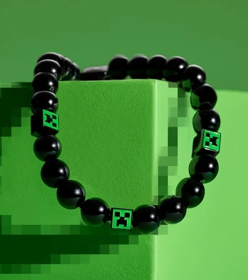 Un bracelet orné de perles rondes en verre noir et de blocs verts gravés au laser avec le visage d’un creeper de Minecraft. Le bracelet repose sur un bloc vert vif inspiré du paysage de Minecraft.