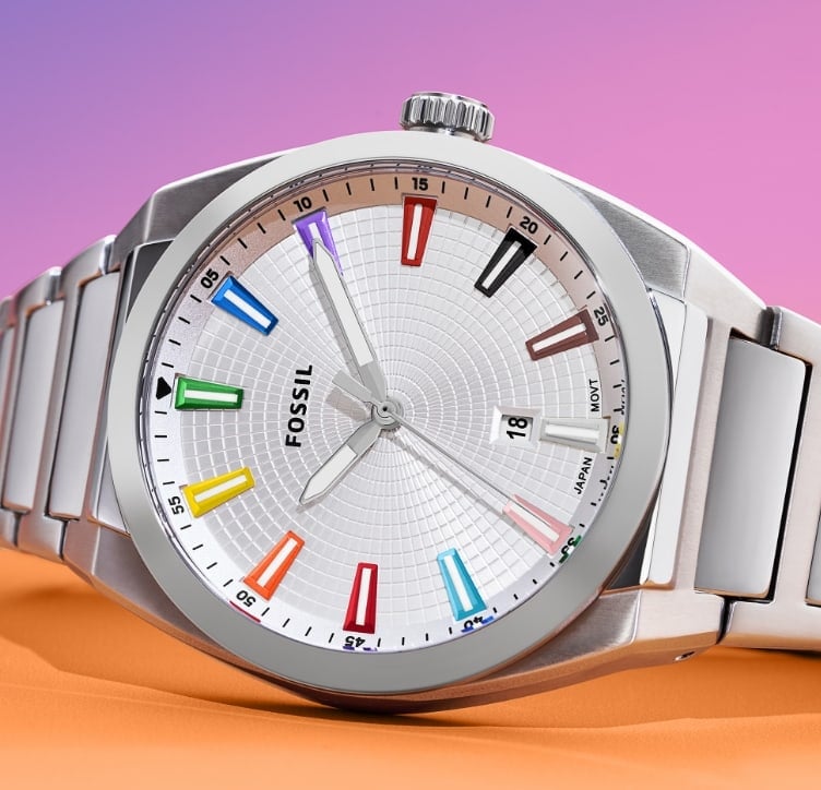 A silver gender-neutral watch featuring a textured dial and rainbow indices that represent the colors of the Pride and Trans flags. The watch is set against a rainbow gradient background that transitions from pink to orange.