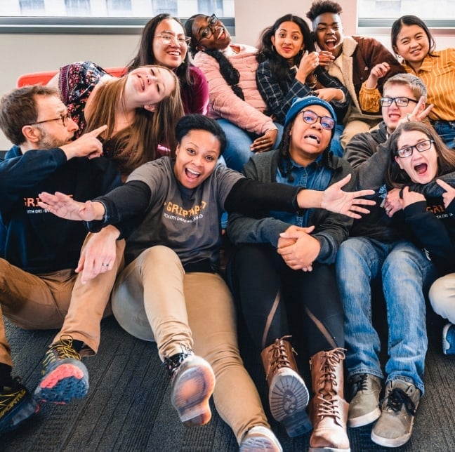 A large group of children sitting on the floor of a classroom smiling and hugging one another..