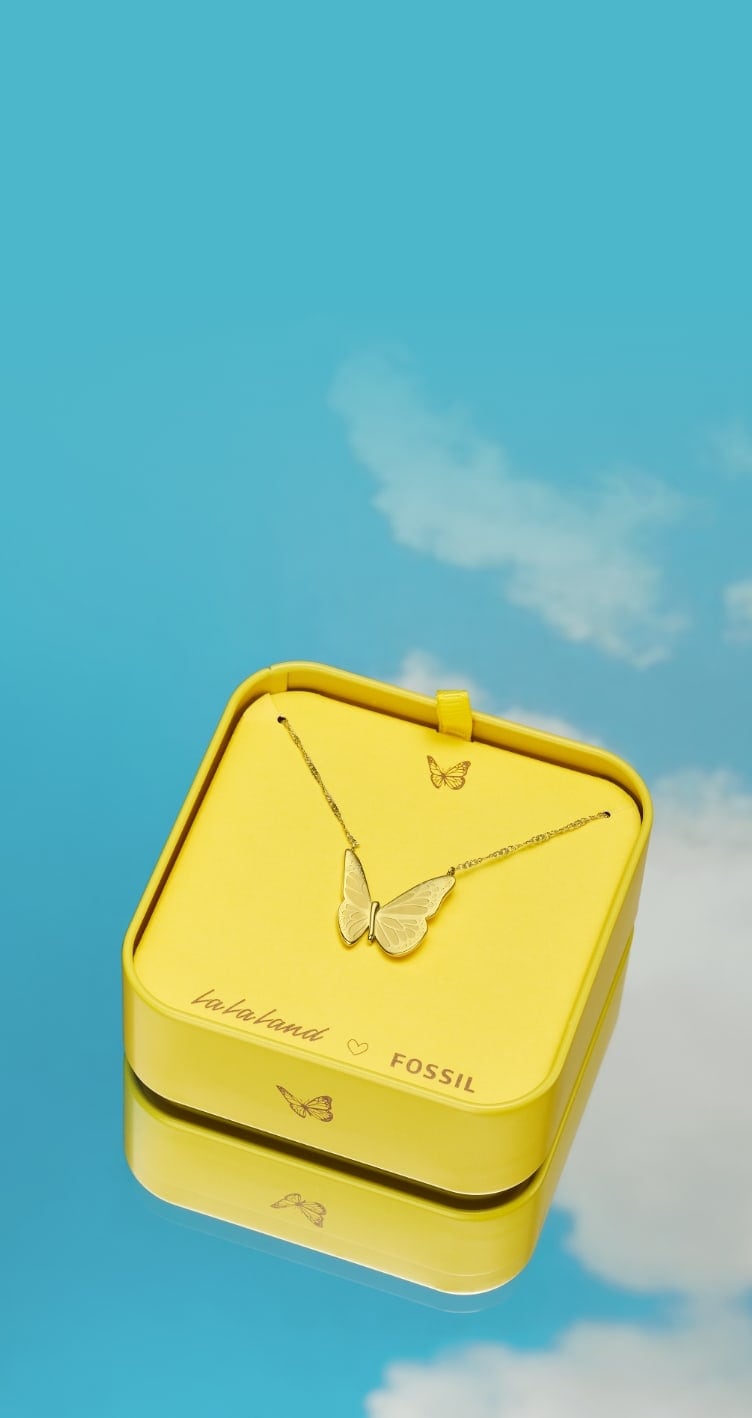 A blue sky and cloud background with a yellow butterfly graphic. Yellow La La Land x Fossil tin with a gold-tone butterfly necklace inside.