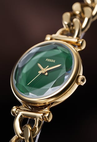 A gold-toned Carlie watch with a green dial sits on the wrist of a stylish woman in a white blouse and jeans.