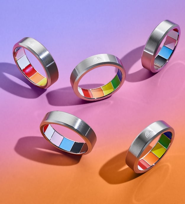 Five silver rings spin upright on their side, displaying a rainbow enamel interior that represents the colors of the Pride and Trans flags. The rings are set on a rainbow gradient background that transitions from light purple to pink to orange.