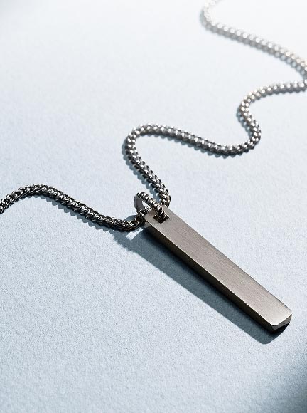 The stainless steel Fossil Ellis ID Necklace, shown in a lifestyle image of a man carrying a surfboard. In an accompanying detail shot, the personalised ID pendant reveals the date 06.21.26.