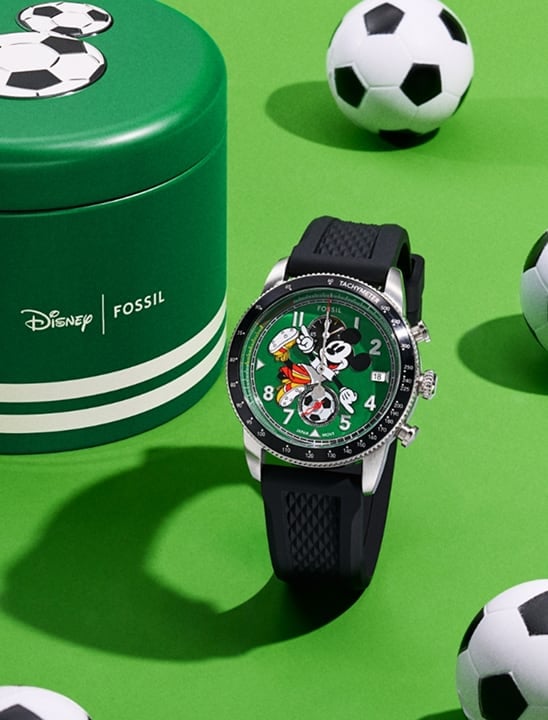 Image of Fossil x Mickey Mouse Soccer watch beside a round watch box on a green background with tiny soccer balls.