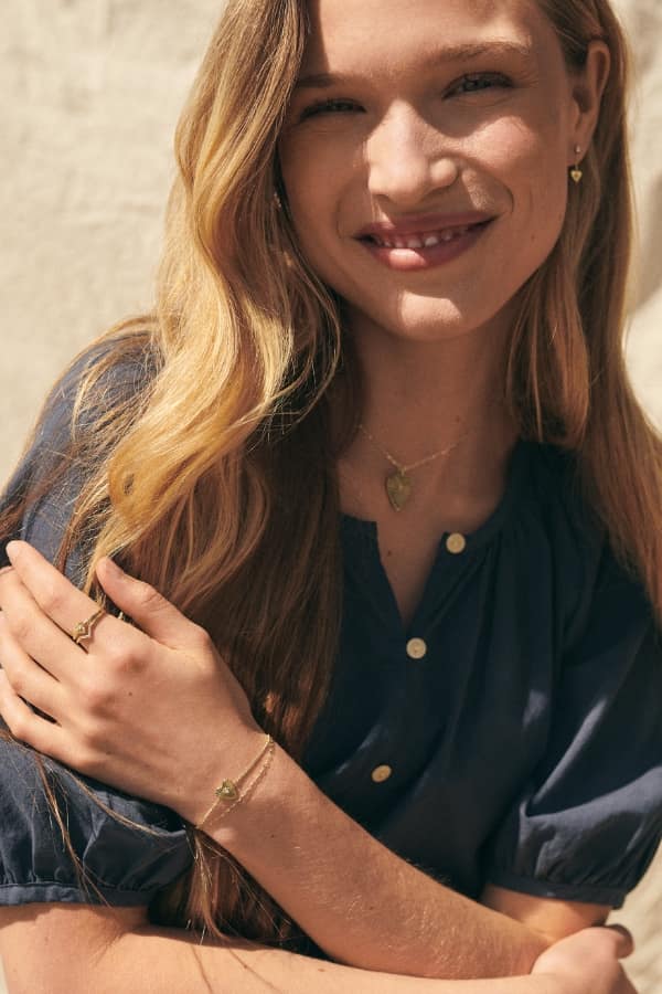 Une femme souriante vêtue d’un chemisier à boutons gris foncé. Elle porte des boucles d’oreilles, un collier et une bague dorés, avec des motifs en émail rose clair.