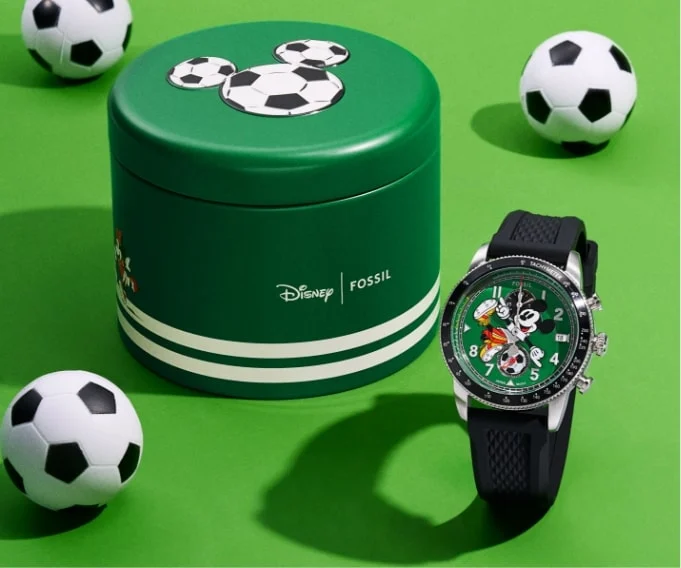 A green cylindrical tin with white stripes is displayed with the Limited-Edition Mickey Mouse Soccer Watch along with mini footballs on a green background.