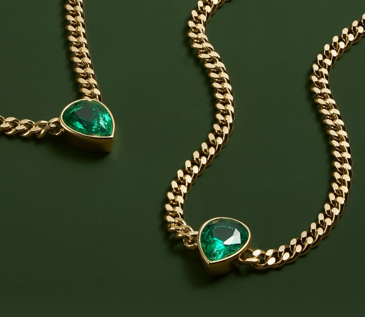A polished brass chain link necklace and bracelet are each accented with an emerald green teardrop crystal. They’re artfully composed on a dark green background.