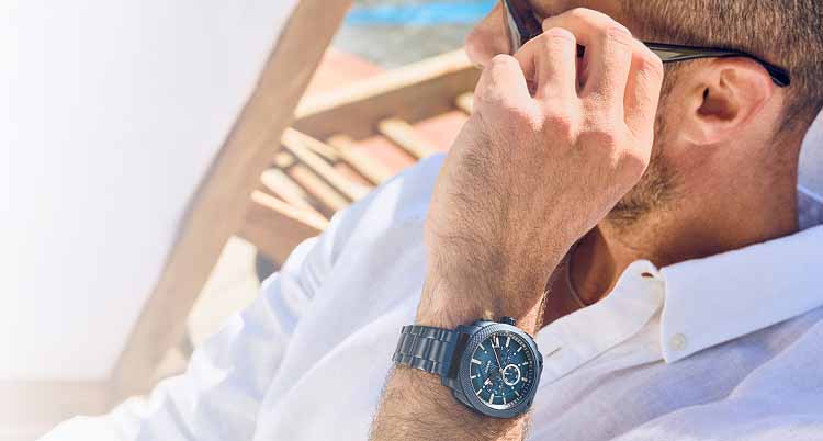 A man sitting poolside, adjusting his sunglasses. Dressed in a white button-down shirt, he's wearing the Fossil Machine Chronograph watch in ocean blue.