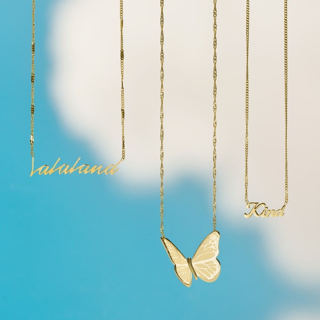 Yellow butterfly graphics. Three gold-tone La La Land x Fossil necklaces on a blue sky background with clouds. The necklaces feature the word LaLaLand, a butterfly motif and the word Kind.