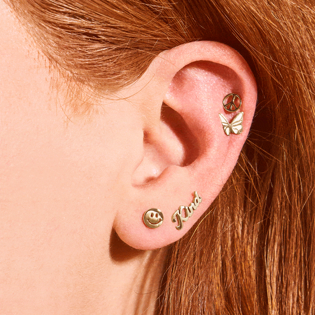 A GIF of a woman's ear showing multiple ways to wear the gold-tone La La Land x Fossil stud earrings, including a peace sign, a smiley face, a butterfly and the word Kind.