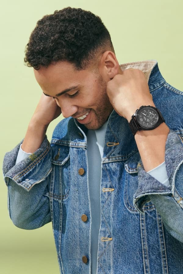 A man in a denim jacket with the collar popped, wearing a sporty, oversized black watch. 