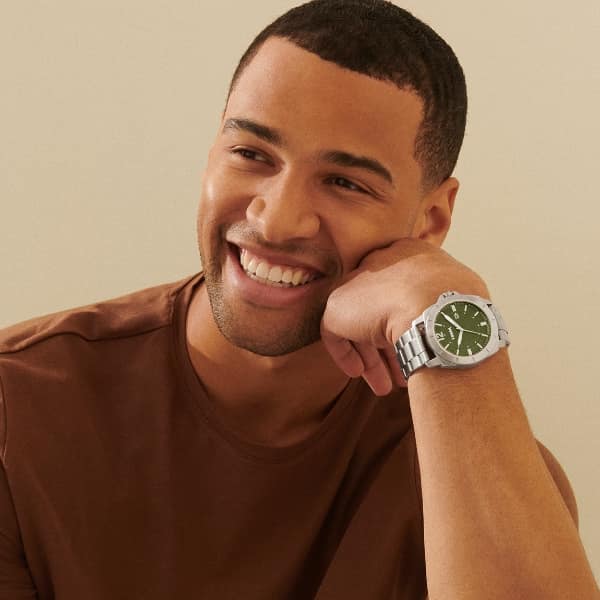 A man is smiling and resting his face on his hand. He’s wearing a brown t-shirt and classic silver men’s watch with an olive-green dial. 