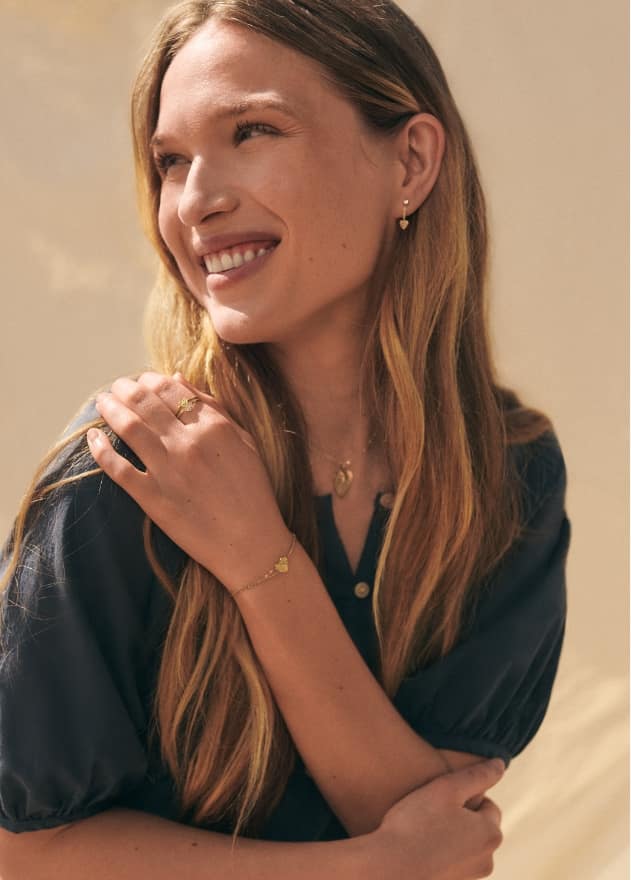 Ein Model, das lächelnd in die Ferne blickt und seine Hand auf seiner Schulter hat. Es trägt goldfarbene Ohrringe, eine Halskette und einen Ring, alle mit hellrosafarbenen Emaille-Motiven.