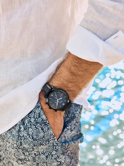 A man stands casually with his hands in the pockets of his navy blue and white floral board shorts, showcasing the midcentury Fossil Neutra Chronograph watch in a deep blue.