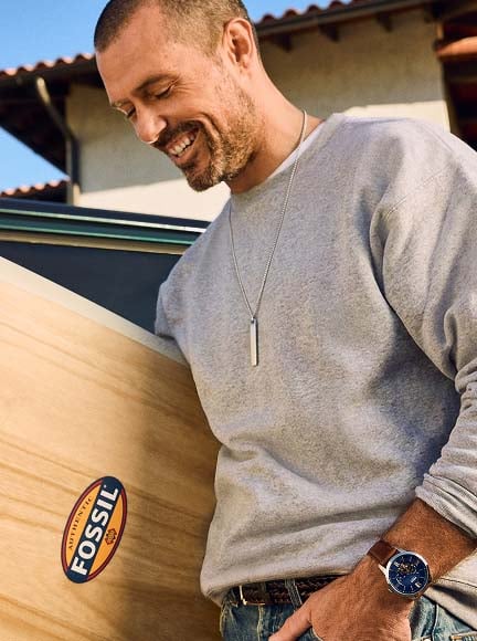 The stainless steel Fossil Ellis ID Necklace, shown in a lifestyle image of a man carrying a surfboard.