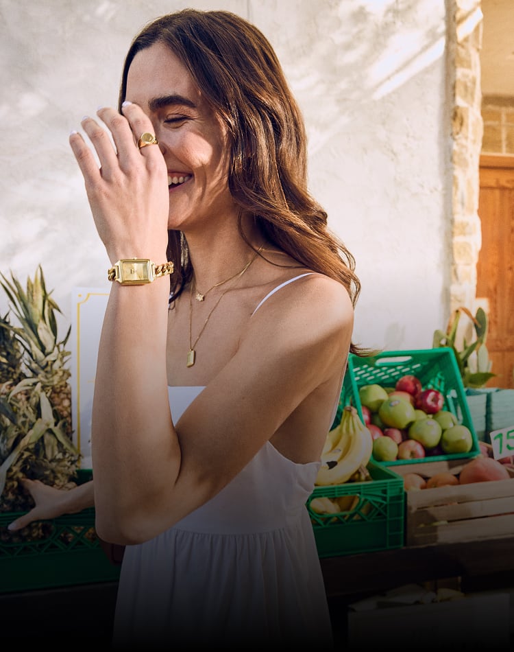 A woman wearing the Fossil Raquel Mini watch and a breezy white sundress, smiling as she walks through a fruit market.
