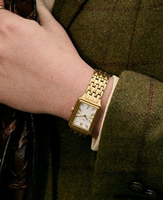 A woman's hand clutching a pebbled leather bag in ochre, wearing a Raquel watch with a gold-tone finish and black dial.