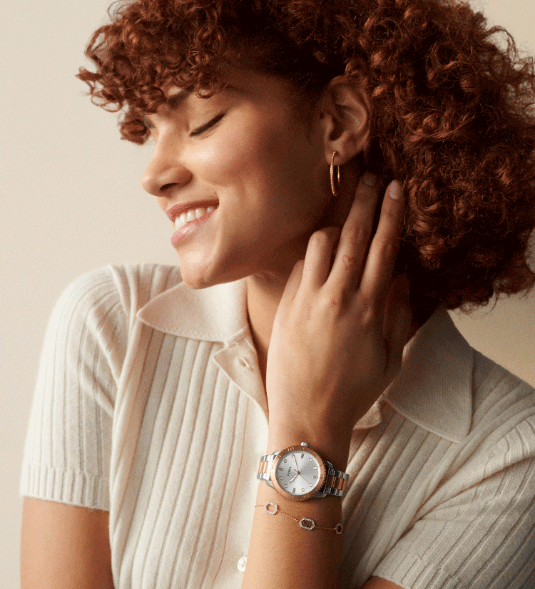 A woman closing her eyes, smiling with her hand touching her neck. She’s wearing a cream-coloured ribbed polo shirt, gold-tone hoop earrings, a rose gold-tone and silver watch and rose gold-tone bracelet accented with sparkling, hexagon shapes. A close-up shot of a women’s dive-inspired watch in rose gold-tone with a white dial.