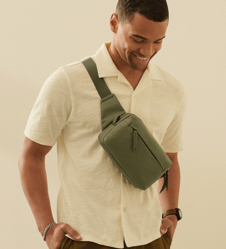 A woman casually sitting on a yellow sofa. She’s wearing a beige short sleeve jumper and trousers, holding a small light blue crossbody handbag. A man smiling and looking downward with his hands in his pockets. He’s wearing a light yellow polo shirt and olive green canvas belt bag worn diagonally across his chest.