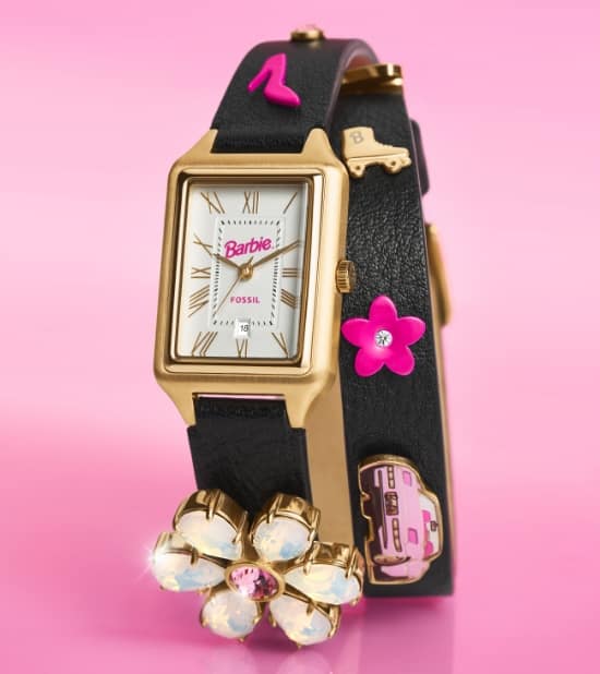 Eine Uhr Barbie x Fossil mit rechteckigem, weißem, satiniertem Zifferblatt mit 1990er-Barbie-Logo, 3-Zeiger-Werk und schwarzem Wickelband aus Leder mit verschiedenen Barbie-Charmanhängern.