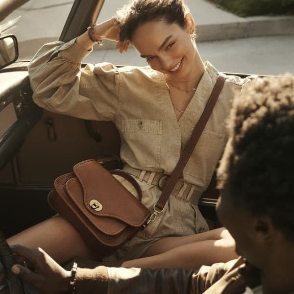 Une femme souriante assise dans une voiture et portant le sac à main Fossil Heritage en cuir brun.