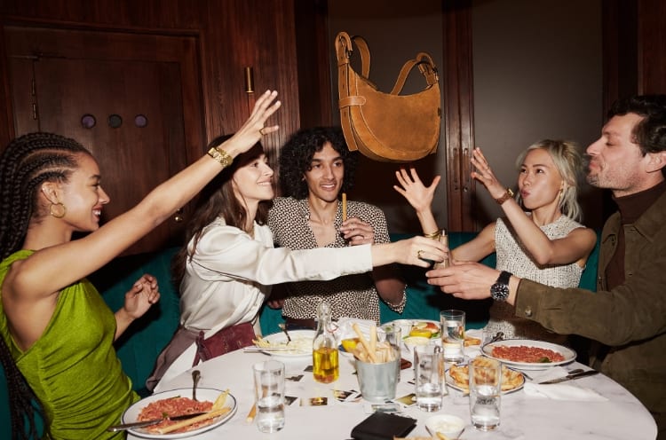 Cinq amis attablés pour le dîner, lançant en l’air un sac Harwell en velours marron. 