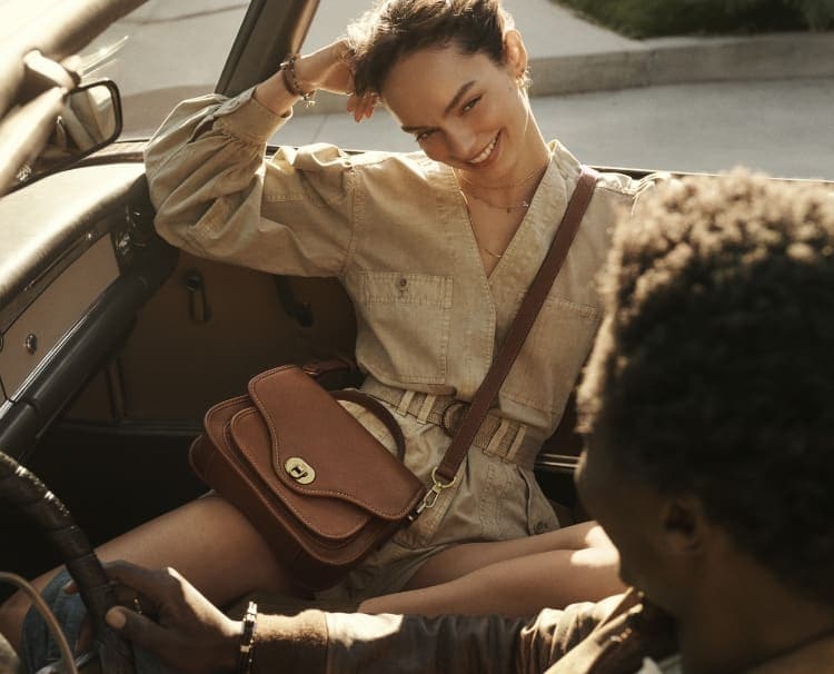 A woman and man smiling in a car and wearing the brown leather Fossil Heritage handbag.