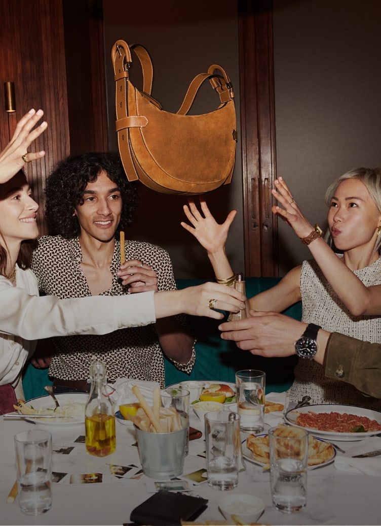 A group of friends sitting around a dinner table and throwing a brown suede Harwell bag into the air.