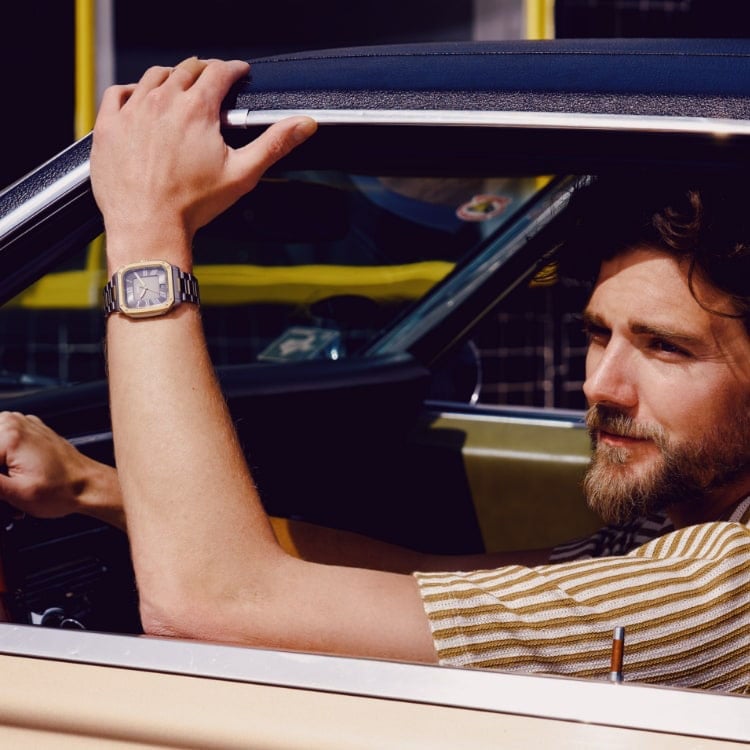 A man in the driver's seat of a vintage car, looking out the window. He's wearing the Crosby square-case watch and a striped button-down short-sleeved shirt.