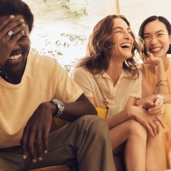 Ein Mann und zwei Frauen, die lachend beieinander sitzen. Sie tragen Produkte aus der Fossil Sommerkollektion. 