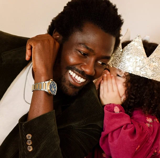 A man smiles at the dinner table as his daughter leans over to whisper something in his ear. He wears a gold-toned Fossil watch and she wears a festive paper crown on her head.