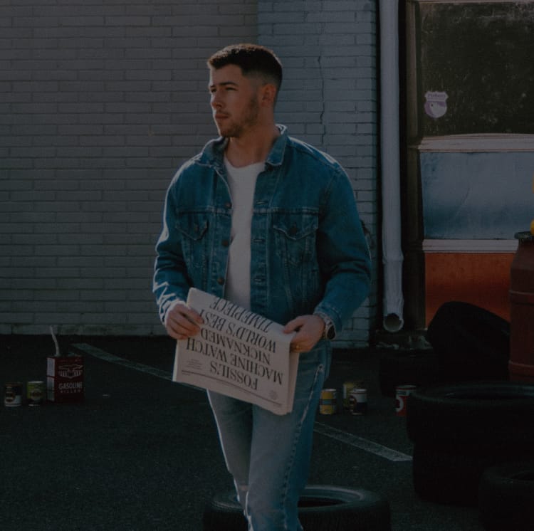 Nick Jonas holding a newspaper and standing near a vintage car at an old-school gas station.